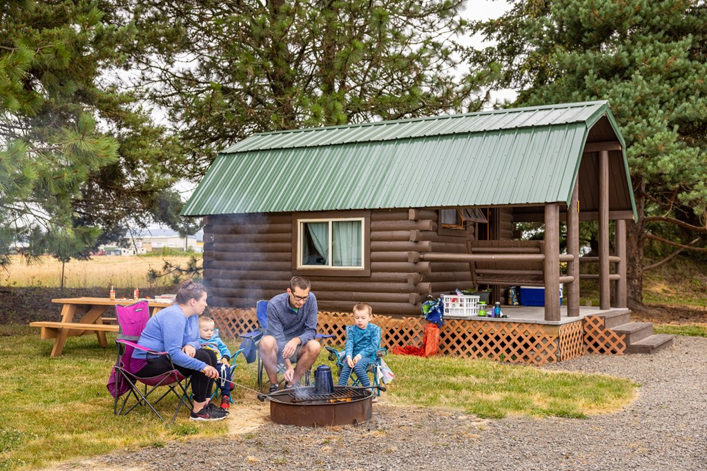 A family roasts marshmallows at a firepit in front of a small log Camping Cabin at a KOA campground.